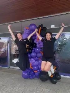 Two women celebrating outside Anytime Fitness at Diamond Creek Shopping with purple balloon decorati.
