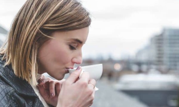 Young woman sipping coffee outdoors near the water with city buildings in the background.